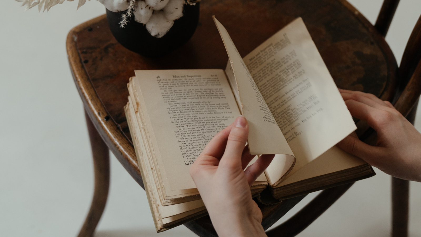 a book on a chair with a dried plant near by. Hands are turning the book pages.