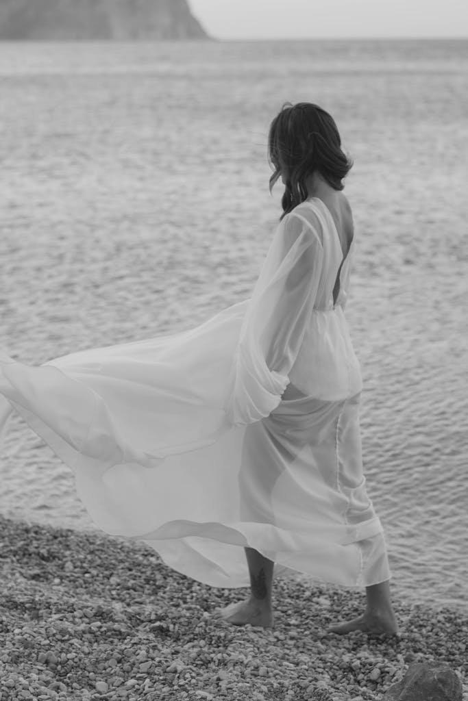 Black and white photo of a woman in a flowing dress by the sea, exuding elegance and serenity.